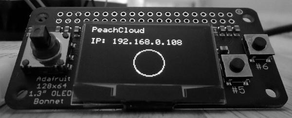 Close-up, black-and-white photo of an Adafruit 128x64 1.3" OLED Bonnet. The circuit board features a 5-way joystick on the left side, two push-buttons on the right side (labelled #5 and #6), and a central OLED display. The display shows text reading: "PeachCloud" on the first line and "IP: 192.168.0.8" on the third line. A circle is displayed beneath the two lines of text and is horizontally-centered".